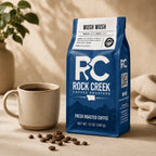 Rock Creek Coffee Roasters Wush Wush coffee bag beside a mug of brewed coffee and scattered roasted beans in warm natural light.