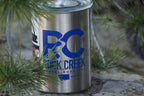 White Rock Creek Coffee Roasters branded metal coffee can with a bold blue RC Rock Creek Coffee Roasters logo visible on the side, surrounded by green pine branches and resting on a stone surface outdoors.