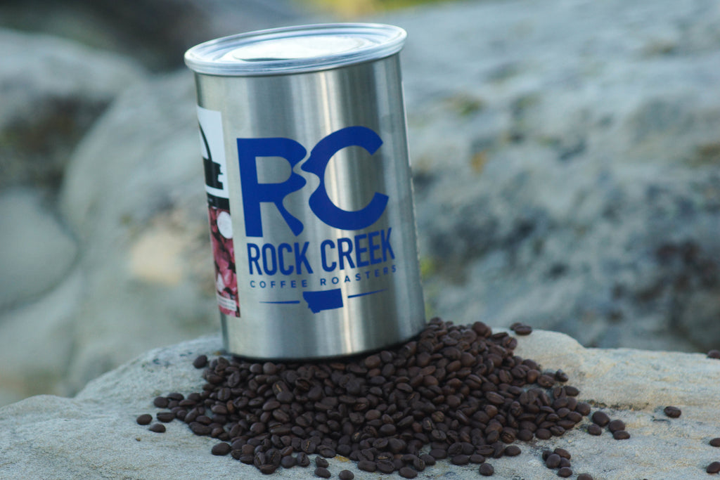 White Rock Creek Coffee Roasters branded metal coffee can with a bold blue RC Rock Creek Coffee Roasters logo on the front, tilted slightly and resting on a pile of roasted coffee beans atop a rock outdoors, with a blurred natural rock background.