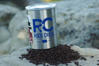 White Rock Creek Coffee Roasters branded metal coffee can with a bold blue RC Rock Creek Coffee Roasters logo on the front, tilted slightly and resting on a pile of roasted coffee beans atop a rock outdoors, with a blurred natural rock background.