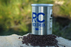 White Rock Creek Coffee Roasters metal coffee can with a bright blue RC logo on the front, sitting upright on a pile of roasted coffee beans atop a natural stone surface outdoors, with green trees and foliage softly blurred in the background.
