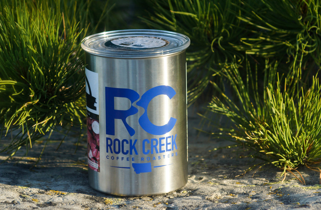 Rock Creek Coffee Roasters metal coffee can with a bold blue RC logo, displayed on a stone surface outdoors and surrounded by green pine branches in natural sunlight.